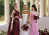 Bride in a white gown stands between two bridesmaids wearing burgundy and blush pink dresses, each holding rich burgundy, wine, blush, and ivory silk wedding bouquets from the Stella Collection. The bridal party smiles together beneath a grand stone colonnade with coordinating floral arrangements lining the walkway.