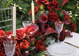 Elegant wedding reception table featuring a lush Stella Collection silk floral runner with deep red roses, burgundy calla lilies, rich red blooms, and dramatic botanical accents. White taper candles, crystal glassware, gold flatware, and layered white plates complete the romantic tablescape.