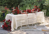 Outdoor wedding reception table styled with the Stella Collection silk floral runner featuring deep red roses, burgundy calla lilies, and rich crimson blooms. White taper candles, crystal Chiavari chairs, gold flatware, and elegant place settings create a dramatic garden-inspired tablescape surrounded by lush greenery.