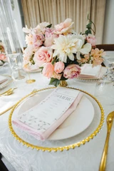 A light pink and ivory floral centerpiece in a gold scalloped vase sits in the center of a rectangle table in front of a place setting featuring a gold beaded rim charger, light pink linen napkin, and gold utensils