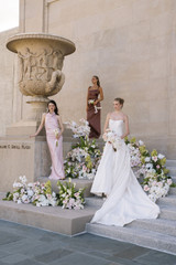 Bride in a modern white gown and two bridesmaids in blush and mocha satin dresses pose on stone steps beside an ornate urn, surrounded by lush floral installations featuring blush anthuriums, white orchids, pale pink roses, and soft green hydrangeas.