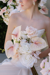 Close-up of a bride in a modern white gown holding a bouquet of blush anthuriums, white roses, and soft ivory blooms, with delicate pearl earrings and romantic florals in the background.