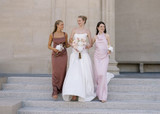 Bride in a modern white gown walks arm-in-arm with bridesmaids in mocha and blush satin dresses on stone steps, carrying bouquets of blush anthuriums, white orchids, and petite white floral posies.