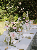 Elegant outdoor reception table featuring airy floral centerpieces of blush anthuriums, white calla lilies, pale pink ranunculus, white orchids, and soft green hydrangeas, styled with crystal glassware and gold flatware.