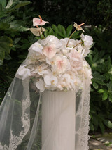 Romantic floral arrangement of blush anthuriums, pale pink roses, white orchids, and soft ivory blooms displayed atop a fluted pedestal and draped with a delicate lace-edged veil.