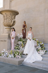 Bride and bridesmaids holding pastel rose bouquets in front of colorful floral ceremony arrangements outdoors.