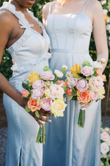 Two bridesmaids in soft blue satin dresses hold colorful bouquets featuring blush and peach roses, pale pink ranunculus, yellow tulips, and fresh greenery.
