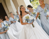 Bride holding a bouquet of blue, white, and green hydrangea while her bridesmaids  wearing light blue satin dresses hold her dress