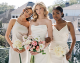 A bride holding a pink and white floral bouquet alongside her bridesmaids in neutral dresses holding small white floral bouquets