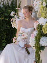 Bride in a sleek white gown holding a cascading bouquet of blush anthuriums, white roses, and soft greenery, seated among lush floral arrangements of pale pink, white, and green blooms.