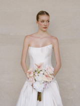 Bride in a strapless white wedding dress holding a bouquet of soft pink and white blooms against a neutral backdrop.