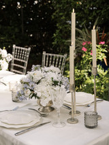 Outdoor wedding reception table featuring a lush white and pale blue floral centerpiece in a vintage silver compote, paired with distressed silver taper candleholders, ivory taper candles, white scalloped dinnerware, crystal glassware, and clear Chiavari chairs surrounded by lush garden greenery.
