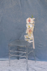 Wedding chair decoration featuring blush anthuriums, cascading white orchids, and white ribbon designed for ceremony aisle seating.