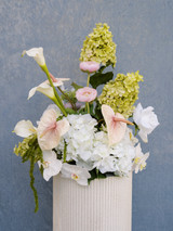 Large vaseless floral arrangement featuring white hydrangeas, blush anthuriums, white orchids, pale pink roses, calla lilies, and soft blush garden roses, styled on a tall ivory pedestal vase against a blue textured backdrop.