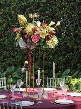 Lush elevated wedding centerpiece featuring deep burgundy calla lilies, ivory calla lilies, green hydrangeas, blush and burgundy garden roses, white peonies, hellebores, Queen Anne's lace, eucalyptus, and cascading burgundy amaranthus arranged on a tall gold stand above a reception table with burgundy linens, gold taper candles, crystal glassware, and clear Chiavari chairs.