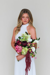 Bride wearing a modern white halter gown holding the Stella Blended Bridal Bouquet, featuring deep burgundy peonies and roses, vibrant pink peonies, white calla lilies, green cymbidium orchids, pale green hydrangea, hellebores, and cascading burgundy amaranthus.