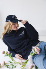 Woman wearing a navy Bouquet Club mockneck sweatshirt and matching navy Bouquet Club baseball cap while reclining with pink flowers.