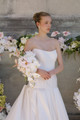 Bride holding a cascading bouquet of white orchids, blush anthuriums, ivory roses, and pale pink flowers in front of a floral display.