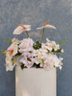 Wedding pedestal arrangement featuring blush anthuriums, white orchids, pale pink roses, and soft garden blooms displayed on a textured column.