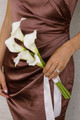 Bridesmaid in a bronze satin dress holding the White Calla Lily Petite Bouquet, featuring four white calla lilies with long stems and flowing ivory ribbon.