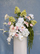 Large ceremony arrangement featuring white hydrangeas, blush anthuriums, white orchids, pale pink roses, calla lilies, and soft blush garden roses styled on a tall ivory pedestal vase against a blue textured backdrop.