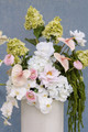 Large ceremony arrangement featuring white hydrangeas, blush anthuriums, white orchids, pale pink roses, calla lilies, and soft blush garden roses styled on a tall ivory pedestal vase against a blue textured backdrop.