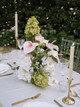 Floral arrangement featuring white hydrangeas, blush anthuriums, white orchids, pale pink roses, calla lilies, and soft blush garden roses, styled in the middle of a wedding reception table with gold candlesticks.