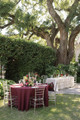 Outdoor garden wedding reception featuring elegant tablescapes beneath towering oak trees. Round tables are dressed in rich burgundy and white linens with clear Chiavari chairs, gold flatware, taper candles, crystal glassware, and lush faux floral centerpieces in burgundy, blush, white, and soft green, creating a sophisticated al fresco wedding setting.