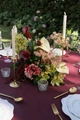 Floral centerpiece featuring deep burgundy and white anthurium, green cymbidium orchids, pale green hydrangea, blush pink peonies, airy pink peonies, and cascading burgundy amaranthus arranged on a burgundy linen. The outdoor wedding tablescape is accented with antique gold taper candleholders, textured glass votives, gold flatware, and crystal glassware, creating a sophisticated garden reception setting.