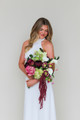 Bride wearing a modern white halter gown holding the Stella Blended Bridal Bouquet, featuring deep burgundy peonies and roses, vibrant pink peonies, white calla lilies, green cymbidium orchids, pale green hydrangea, hellebores, and cascading burgundy amaranthus.