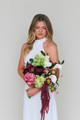 Bride wearing a modern white halter gown holding the Stella Blended Bridal Bouquet, featuring deep burgundy peonies and roses, vibrant pink peonies, white calla lilies, green cymbidium orchids, pale green hydrangea, hellebores, and cascading burgundy amaranthus.