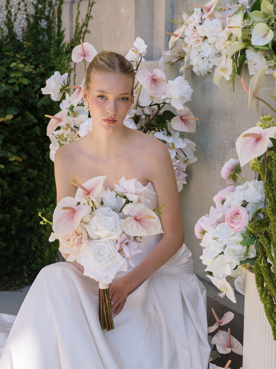 Bride holding wedding bouquet featuring blush pink anthurium, roses, and ivory flowers held in front of a bridal gown.