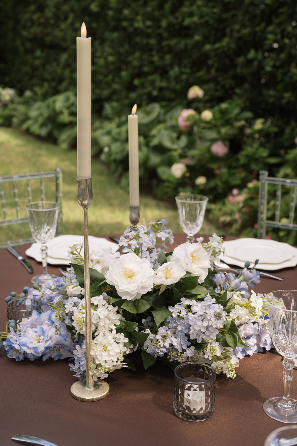 wedding reception table featuring a lush white and pale blue floral meadow centerpiece with white garden roses, hydrangeas, and delicate blue blossoms, accented by distressed silver taper candleholders, crystal stemware, white dinnerware, and clear Chiavari chairs against a lush garden backdrop.