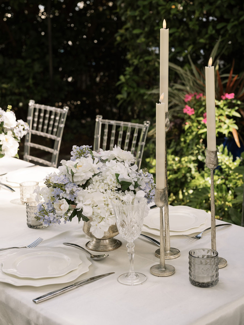 Outdoor wedding reception table featuring a lush white and pale blue floral centerpiece in a vintage silver compote, paired with distressed silver taper candleholders, ivory taper candles, white scalloped dinnerware, crystal glassware, and clear Chiavari chairs surrounded by lush garden greenery.