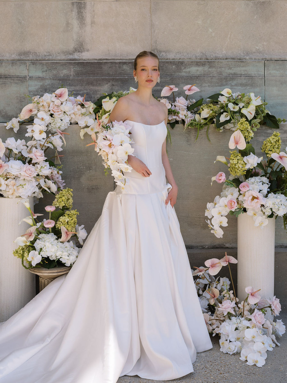 Bride in a white wedding gown standing among lush floral arrangements featuring white orchids, blush anthuriums, roses, and hydrangeas.