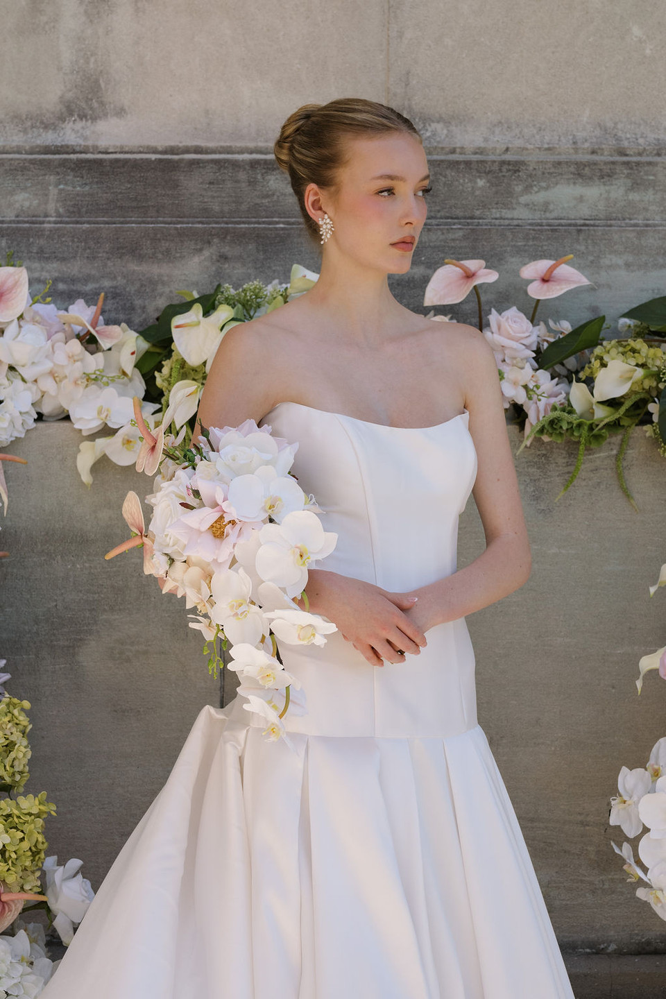 Bride holding a cascading bouquet of white orchids, blush anthuriums, ivory roses, and pale pink flowers in front of a floral display.
