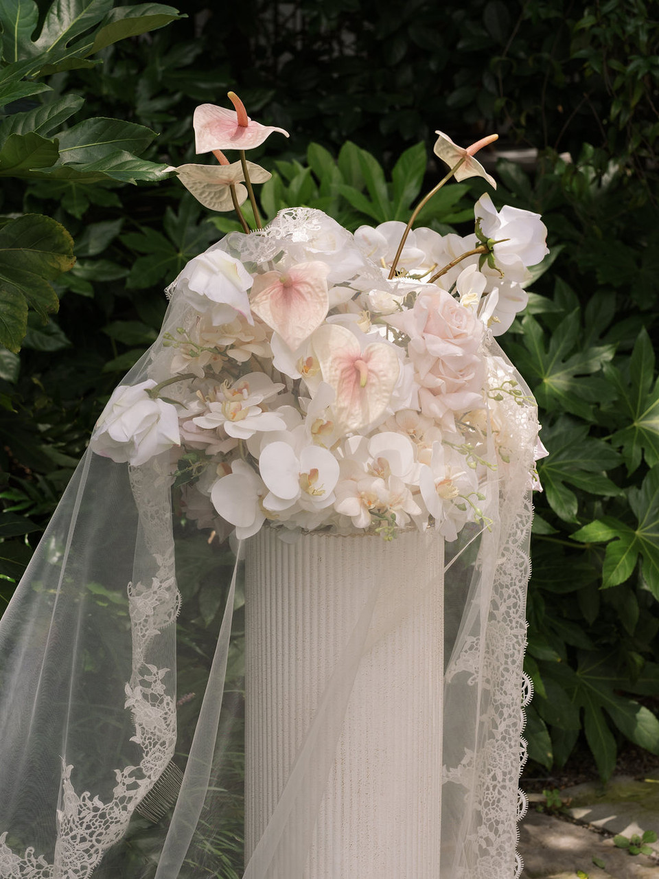 Large pedestal floral arrangement with blush anthuriums, orchids, and roses styled with a lace bridal veil.