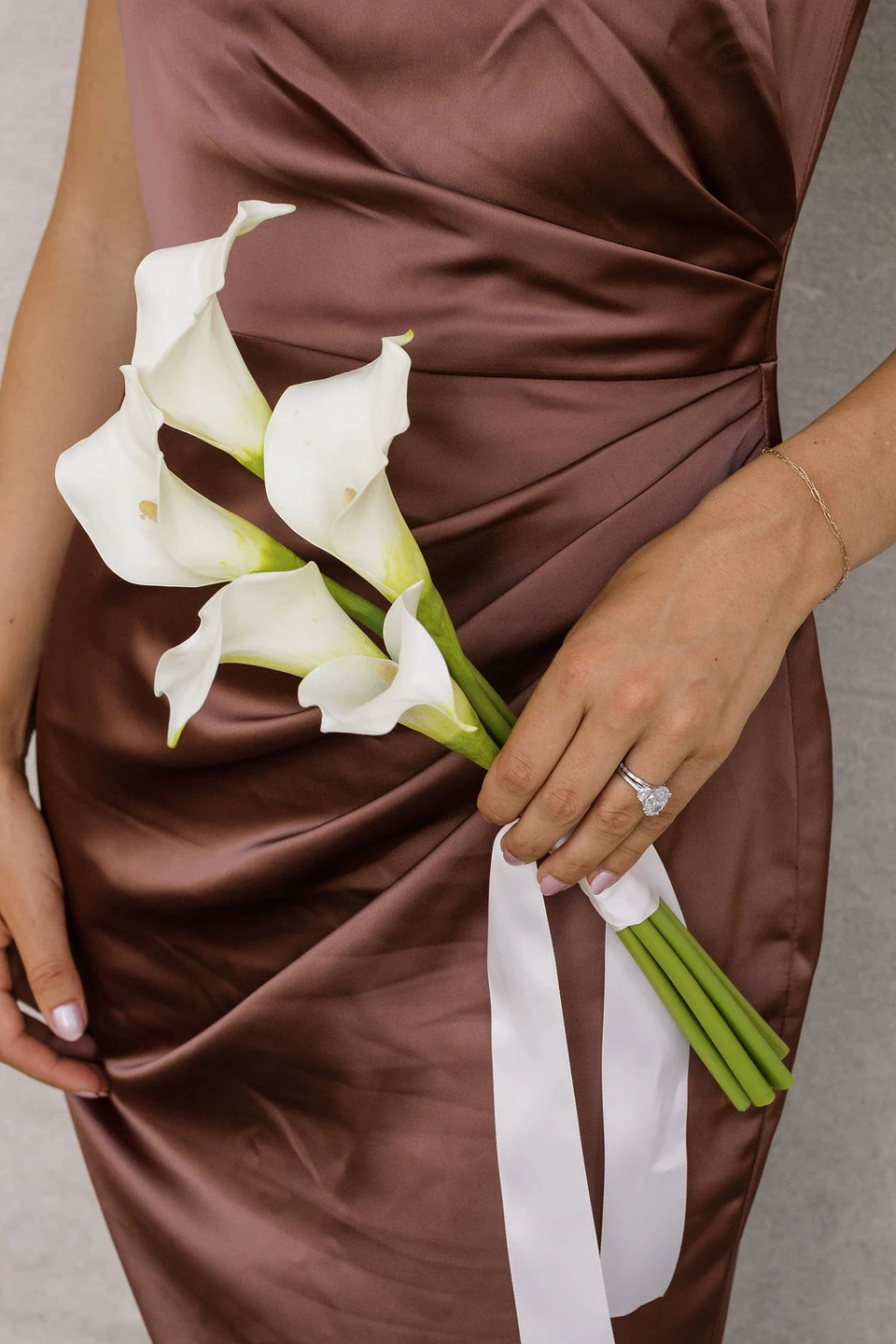 Bridesmaid in a bronze satin dress holding the White Calla Lily Petite Bouquet, featuring four white calla lilies with long stems and flowing ivory ribbon.