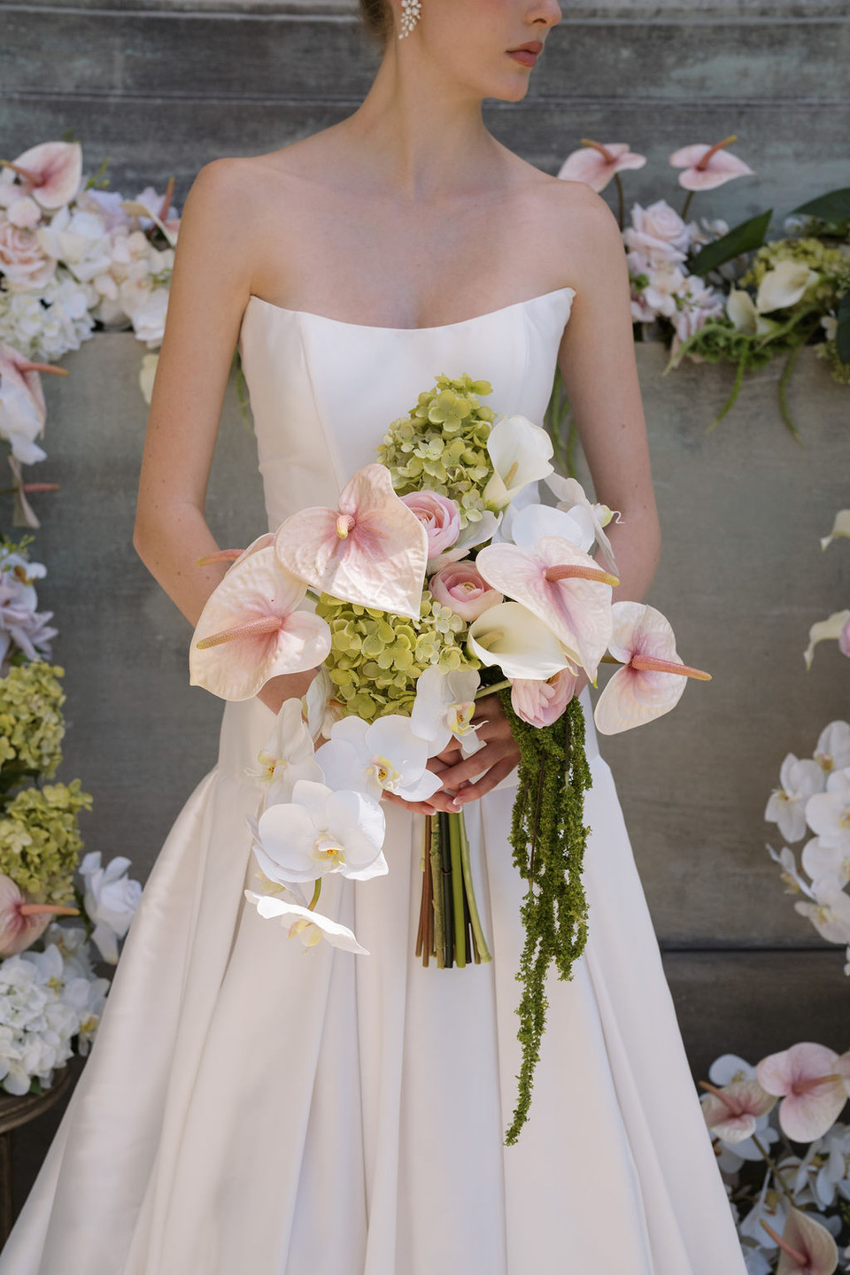 Bride holding a cascading bouquet with blush anthuriums, orchids, hydrangeas, and trailing amaranthus in front of a floral installation.
