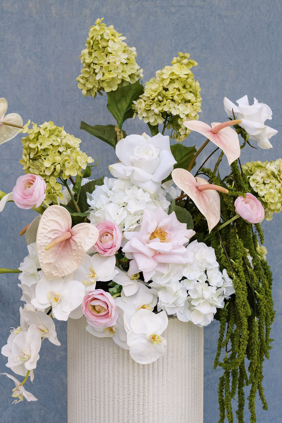 Large ceremony arrangement featuring white hydrangeas, blush anthuriums, white orchids, pale pink roses, calla lilies, and soft blush garden roses styled on a tall ivory pedestal vase against a blue textured backdrop.