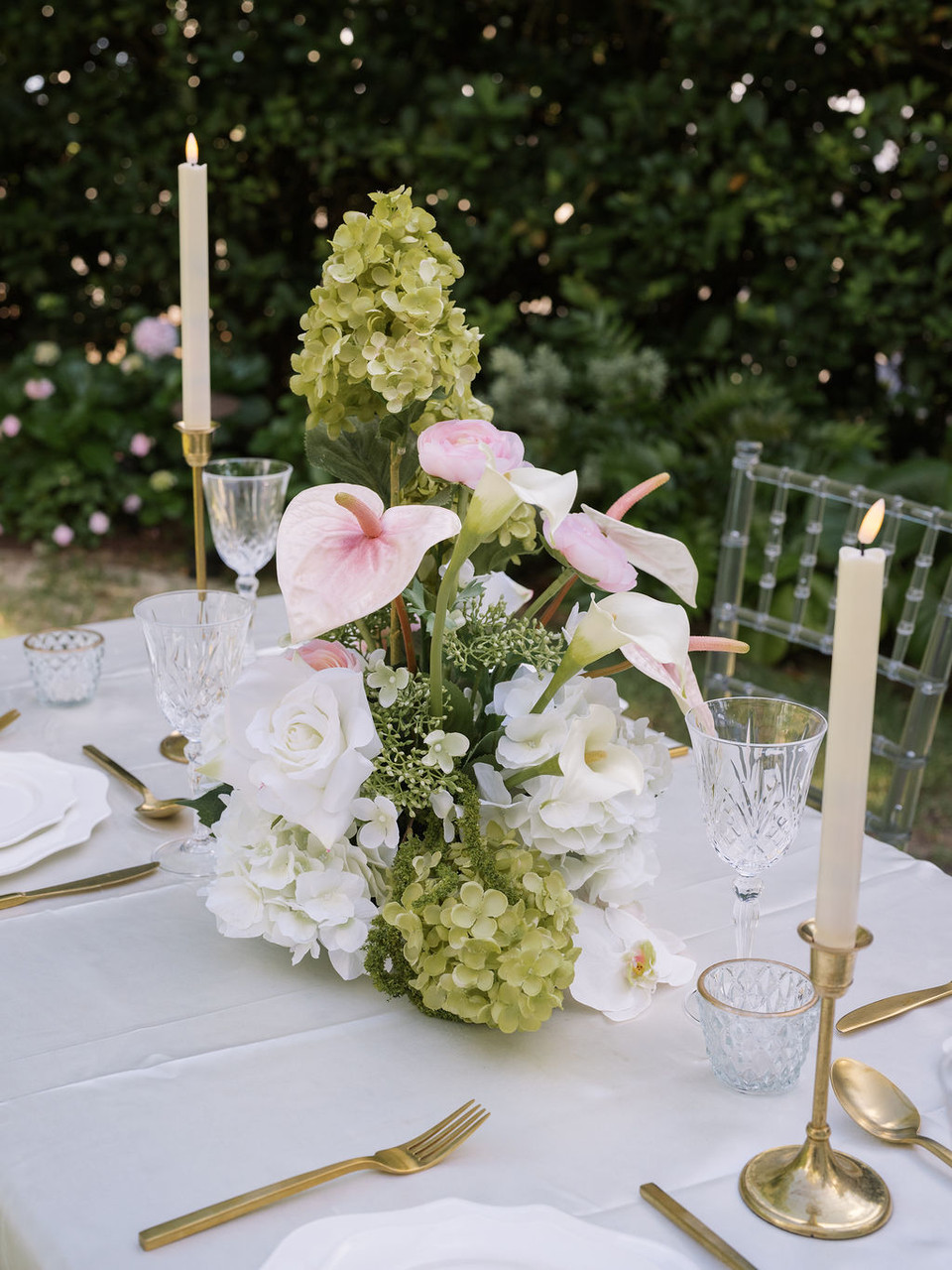 Floral arrangement featuring white hydrangeas, blush anthuriums, white orchids, pale pink roses, calla lilies, and soft blush garden roses, styled in the middle of a wedding reception table with gold candlesticks.