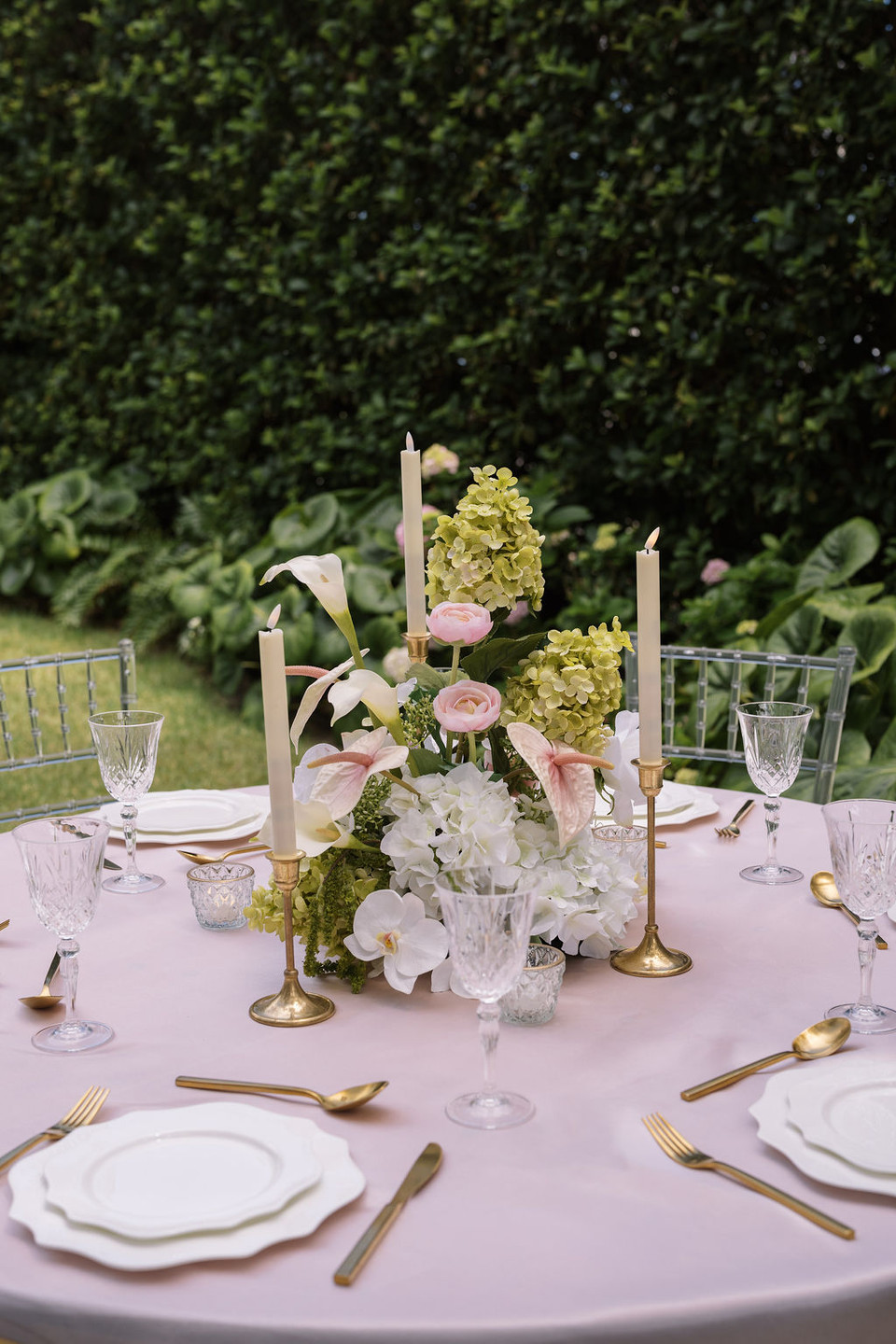 Floral arrangement featuring white hydrangeas, blush anthuriums, white orchids, pale pink roses, calla lilies, and soft blush garden roses, styled in the middle of a wedding reception table with gold candlesticks.
