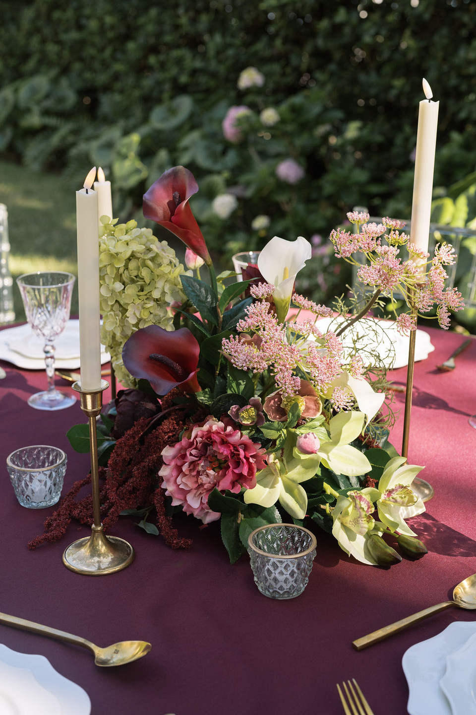 Floral centerpiece featuring deep burgundy and white calla lilies, green cymbidium orchids, pale green hydrangea, blush pink peonies, airy pink peonies, and cascading burgundy amaranthus arranged on a burgundy linen. The outdoor wedding tablescape is accented with antique gold taper candleholders, textured glass votives, gold flatware, and crystal glassware, creating a sophisticated garden reception setting.