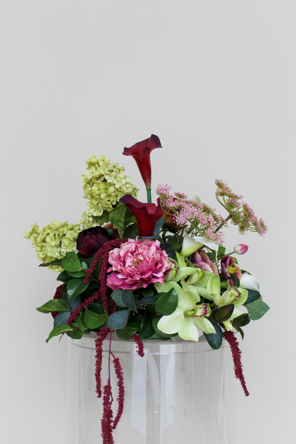 Stella blended large vaseless centerpiece featuring deep burgundy calla lilies, white calla lilies, blush peonies, pale green hydrangeas, green cymbidium orchids, burgundy roses, white anthuriums, cascading burgundy amaranthus, and airy pink accent blooms.