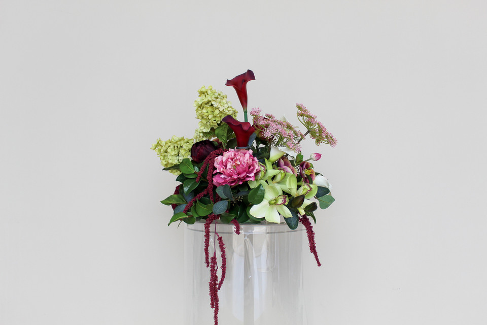 Stella blended large vaseless centerpiece featuring deep burgundy calla lilies, white calla lilies, blush peonies, pale green hydrangeas, green cymbidium orchids, burgundy roses, white anthuriums, cascading burgundy amaranthus, and airy pink accent blooms.