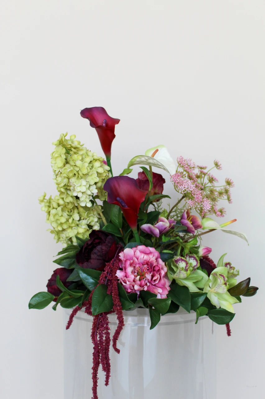 Floral centerpiece featuring deep burgundy and white anthurium, green cymbidium orchids, pale green hydrangea, blush pink peonies, airy pink peonies, and cascading burgundy amaranthus.