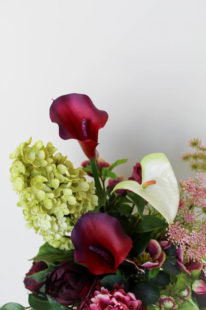 Floral centerpiece featuring deep burgundy and white anthurium, green cymbidium orchids, pale green hydrangea, blush pink peonies, airy pink peonies, and cascading burgundy amaranthus.
