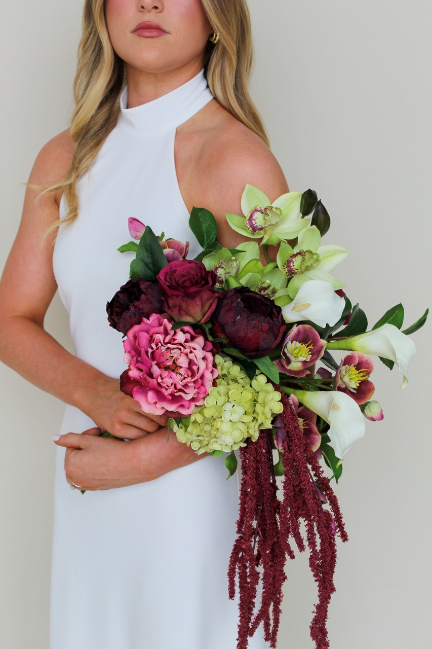 Bride wearing a modern white halter gown holding the Stella Blended Bridal Bouquet, featuring deep burgundy peonies and roses, vibrant pink peonies, white calla lilies, green cymbidium orchids, pale green hydrangea, hellebores, and cascading burgundy amaranthus.