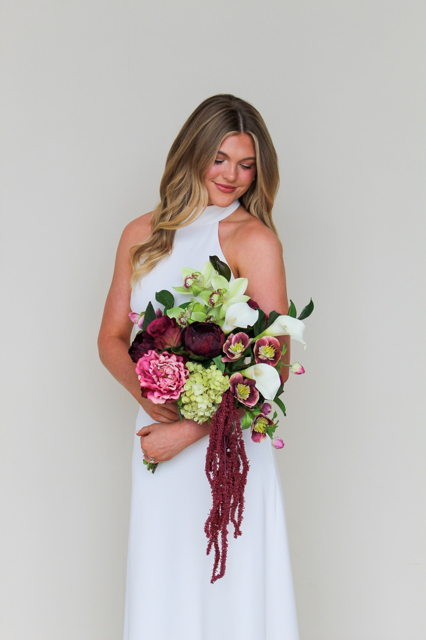 Bride wearing a modern white halter gown holding the Stella Blended Bridal Bouquet, featuring deep burgundy peonies and roses, vibrant pink peonies, white calla lilies, green cymbidium orchids, pale green hydrangea, hellebores, and cascading burgundy amaranthus.