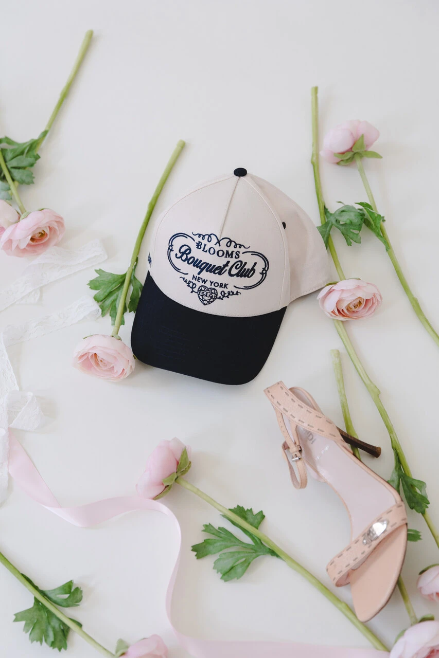 The Bouquet Trucker Club hat laid out on the floor with pink ranunculus stems and a nude heel